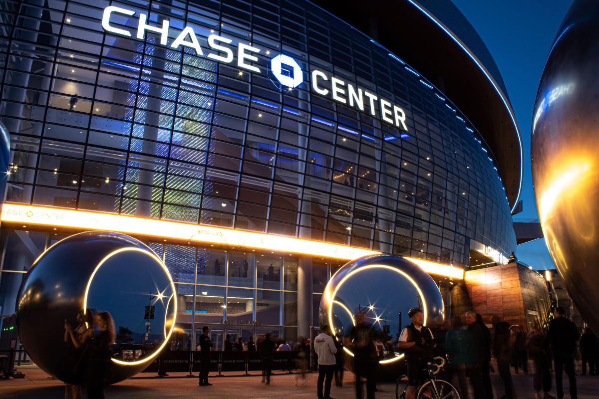 Chase Center arena at dusk, its curved glazed facade lit from within above the entrance canopy